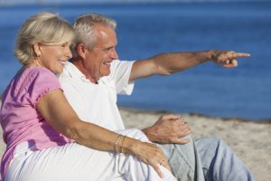 Senior man and woman couple sitting on a deserted beach pointing to the sea