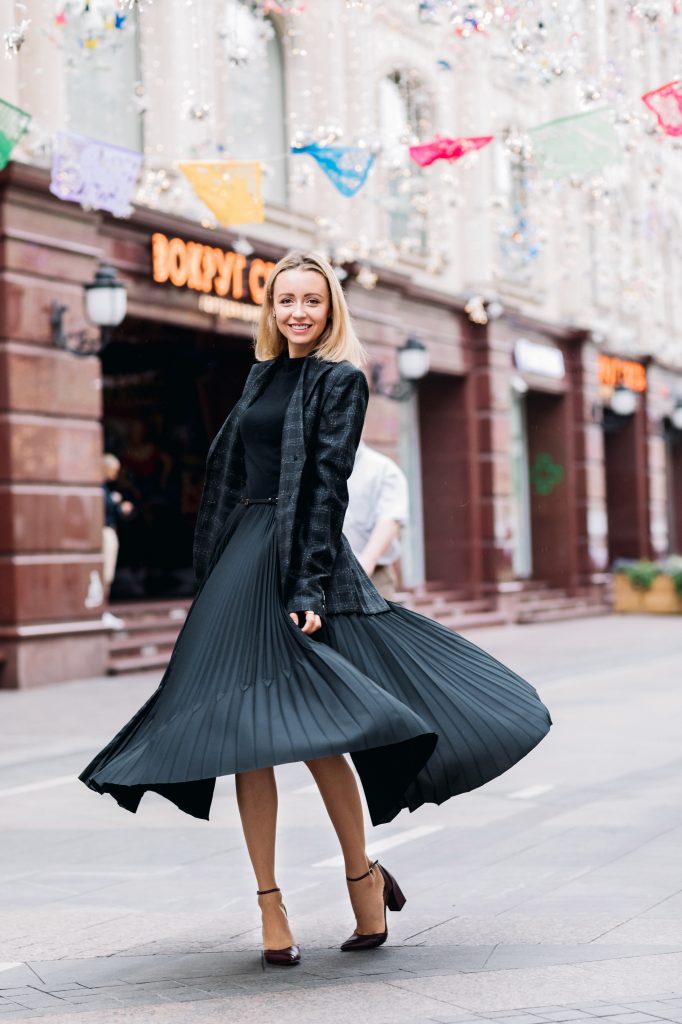 femme blonde portant un jupe plissée midi un pull noir et un blazer foncé à carreaux avec des babies pointues à talons